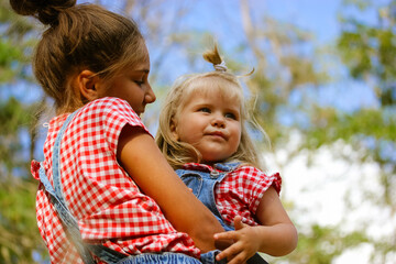 Two happy sisters in summer forest. Older teenage mestizo girl holding baby sister blonde girl 3 years old. Happy family. Related people. Showing love, care, warm feelings. Multiethnic kids have fun.
