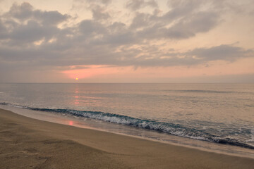 Rising sun and its reflection in sea in early morning. Cloudy sunrise at deserted sandy beach. Slight morning surf waves