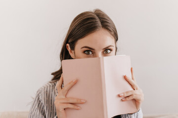 Green-eyed girl in striped pajamas looks into camera and covers her face with notebook. Portrait of...