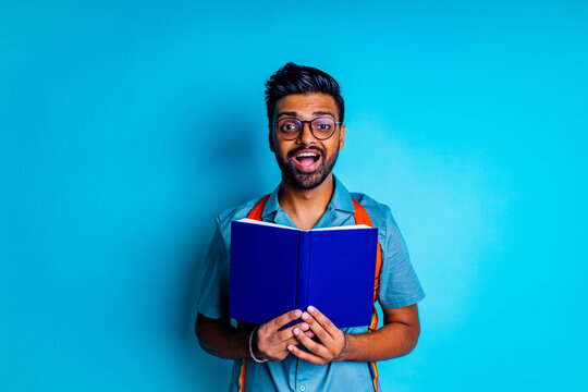 handsome young bearbed indian man with eye glasses in blue cotton t-shirt with orange rainbow backpack in studio background