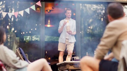 Male house owner standing in doorway, eating nachos and talking to friends sitting by bonfire at summer party. People gathering together again