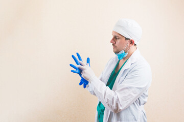 Young doctor in white suit and protective breathing mask puts gloves on his hands on yellow background isolated.