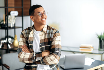 Portrait of an attractive confident positive young mixed race latino man with glasses, designer or freelancer, stylishly casual dressed, stand near a desk with arms crossed, looking to the side, smile