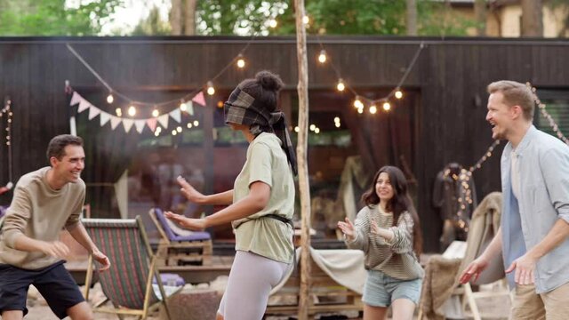 Diverse young friends having fun playing blind man's buff tag game at summer party. Blindfolded woman attempting to touch other players clapping hands around in backyard