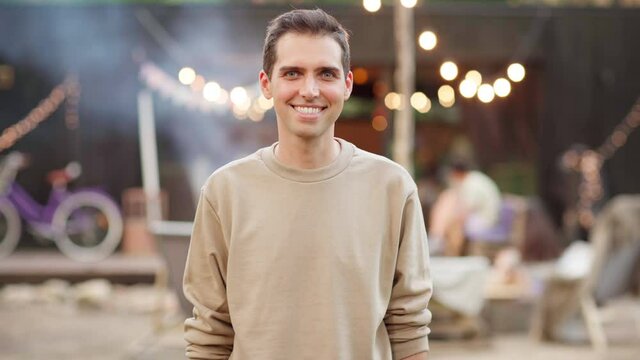 Zoom in portrait of handsome happy young man looking at camera and smiling cheerfully standing against background of outdoor barbecue summer party with smoke and lights