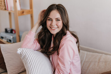 Naklejka premium Portrait photo at home. Beautiful brunette girl dressed in pink sweatshirt brightly smiling and posing. Curly woman resting on sofa in white cozy room