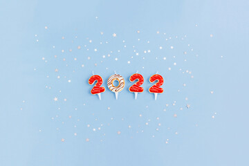 Candles in the form of numbers 2022 on light blue background decorated with silver star confetti. New Year banner