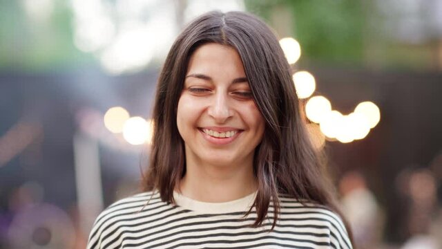 Head and shoulders portrait of authentic young Middle Eastern woman in striped sweatshirt looking at camera, smiling and laughing happily against background of barbecue party with smoke and lights