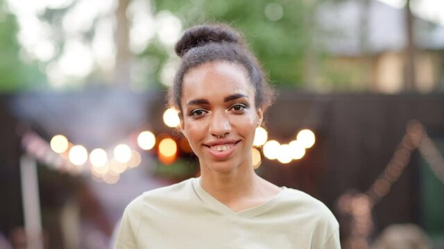 Head and shoulders portrait of beautiful young mixed race Black woman with septum nose piercing looking at camera and smiling happily against background of outdoor barbecue party with smoke and lights
