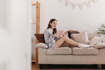 Obraz premium Brunette girl reads book with interest while sitting on soft sofa in her apartment. Woman in pajamas looks surprised. Teen holding notebook and resting at home on sofa