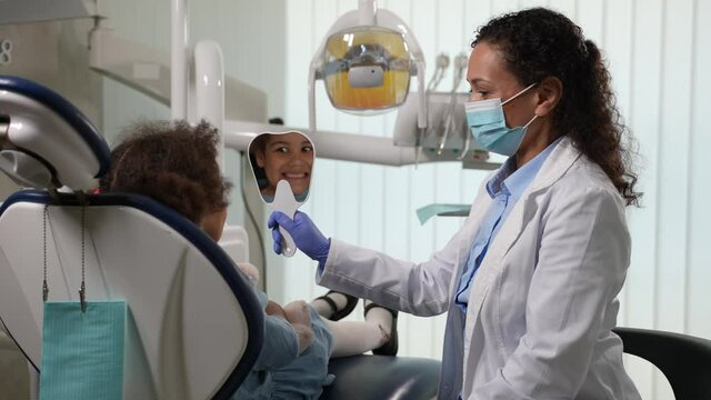 Cute mixed race girl smiling broadly while looking at mirror after teeth treatment. Masked female pediatric stomatologist showing to elementary age child patient her treated teeth in dental office