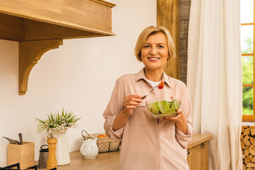 Modern senior aged mature woman eating fresh green salad and vegetables in kitchen, smiling happy....