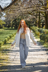Positive caucasian female have fun in park, surrounded by green plants trees, smiling, looking at camera, in motion. Redhead lady in casual stylish clothes outfit, full-length portrait