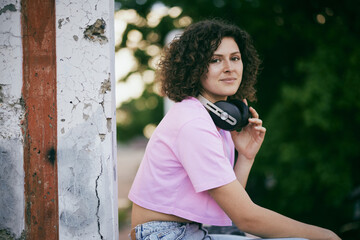 A young attractive woman with curly hair sitting outdoors with headphones in her hands and chilling.