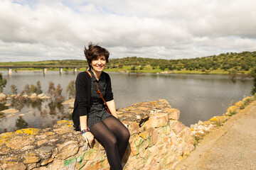 Attractive young female sitting on top of a rock while witnessing the ruins of an old bridge that...