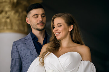 Portrait of young bride and groom indoors. Stylish groom looks at his lovely bride. Beautiful newlyweds are smiling and enjoying their wedding. Bride with elegant hairstyle