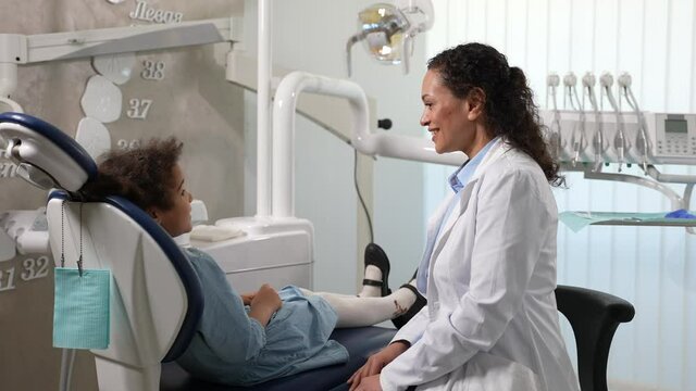 Cute preadolescent mixed race girl and woman stomatologist communicating in denal office before examination. Friendly pediatric dentist giving high five little patient sitting on stomatological chair