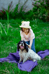 a small, beautiful girl in a hat, combing her dog, in the park on a blanket and green grass