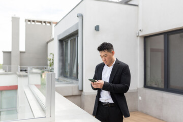 Freelancer Asian businessman in business suit enjoys phone standing on balcony terrace of modern office center