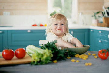 Happy childhood in a family in a country house. A beautiful little child blonde boy of European appearance in the kitchen in the apartment cooks and eats with fresh vegetables.