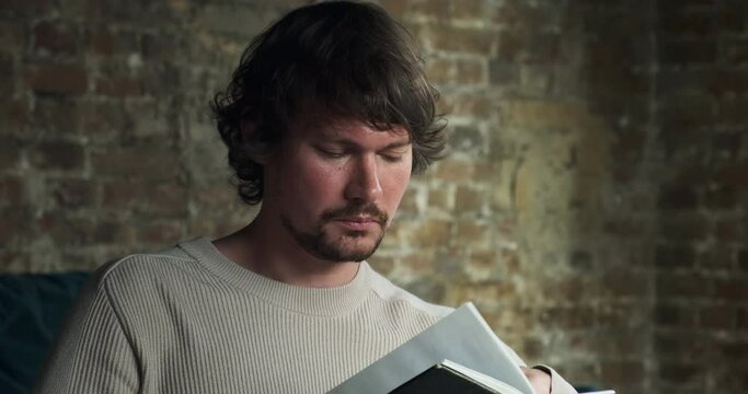 A young bearded man is carefully reading an interesting book, a fiction, a novel, thinking about what he was reading. A quiet day at home without electronic gadgets or mobile phones.