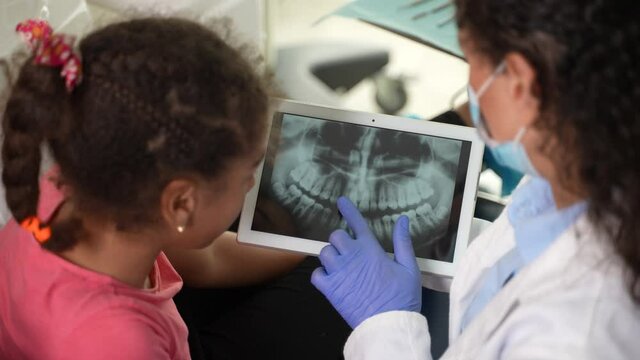 Close-up of tablet with dental x-ray in hands of female dentist, explaining teeth treatment to girl patient. Mixed race preadolescent child and stomatologist examining dental x-ray