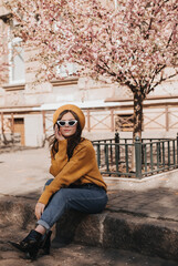 Obraz premium Woman in jeans and sweater is resting on curb against background of sakura. Portrait of lady in stylish outfit in parisian style enjoying spring weather