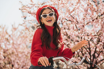 Fototapeta na wymiar Portrait of gorgeous girl in red outfit and sunglasses on background of sakura. Cheerful woman in cashemere sweater and beret smiling and riding bicycle