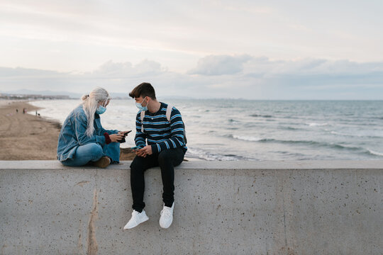 Young couple in masks using smartphones near sea
