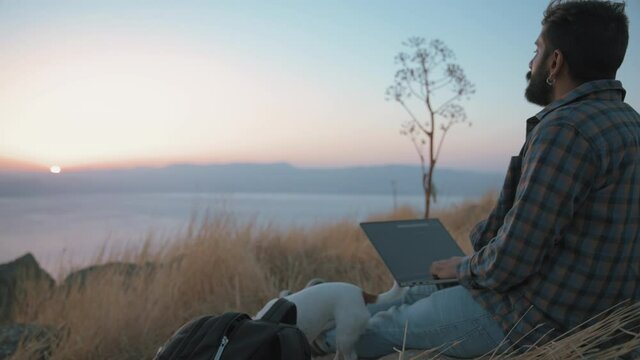 Digital nomad with a laptop and his dog enjoying the sunset. Slow motion. Remote working anywhere, digital nomad concept. 