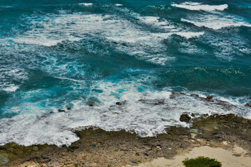Ocean waves are hitting some small rocks at the seashore producing seafoam. High angle view.