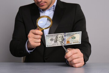 Expert authenticating 100 dollar banknote with magnifying glass at table on light grey background,...