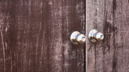 Metal knob on an old door leaf. A pair of stainless steel doorknobs on four dark brown door panels at a corroded surface with copy space. Select focus and subject.