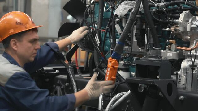 Mechanic is Pressing the Buttons on remote Control. A Mechanic is Lowering Down the Complex Part of the Car engine with Multiple Wires on the Test Bench. A Mechanic is Operating on the Vehicle. 