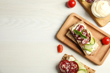 Fresh rye crispbreads with salami, cream cheese and cucumber on white wooden table, flat lay. Space for text