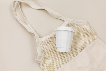 Beige reusable cotton mesh bag and coffee mug on a beige background. Eco-friendly shopping bag....