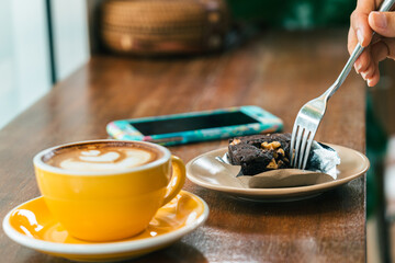 Hands of a young woman with a fork . A plate of cake and smartphone on the table. In the foreground is a cup of coffee