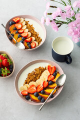 Delicious healthy breakfast, granola with yogurt and sweet fruits, banana and strawberries, blueberries and peaches, milk in a mug and flowers in a vase, top view