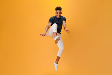 Fototapeta na wymiar Stylish guy emotionally poses and jumps on orange background. Snapshot of handsome dark-haired man in white pants moving on isolated