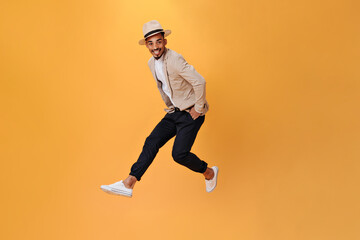 Fototapeta na wymiar Young man in black pants, jacket and hat jumps on orange background. Cheerful guy in suit moving on isolated backdrop