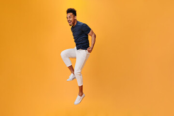 Fototapeta na wymiar Emotional guy in white pants and sneakers jumping on isolated background. Cool man in dark tee posing on orange backdrop