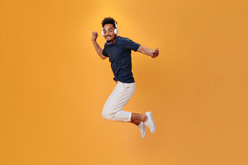 Fototapeta na wymiar Energetic man in stylish outfit jumping on orange background and listening to music on headphones. Cool guy un blue shirt having fun on isolated backdrop