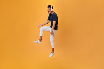 Fototapeta na wymiar Guy in white pants listens to music with headphones and jumps on orange background. Cheerful man in dark blue shirt dancing on isolated backdrop