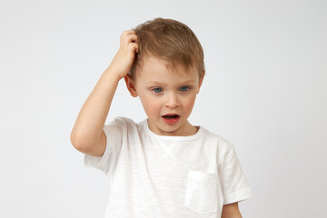 Little boy with blue eyes is deep in thought, looking down and scratching his head against a white background. The child's thought process