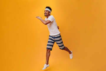 Fototapeta na wymiar Young guy in summer outfit runs on isolated background. Snapshot of man in striped shorts and white tee on orange backdrop