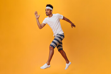 Fototapeta na wymiar Dark-skinned guy in shorts and T-shirt runs on orange background. Sportsman has workout on isolated. Portrait of dark-skinned teen