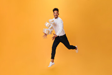 Fototapeta na wymiar Joyful man jumps on orange background and throws up gifts. Dark-haired man in white shirt and black pants happily posing on isolated