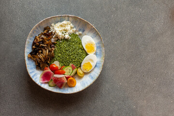 Vegetarian Green rice bowl, kale-almond pesto, oyster and maitake mushrooms, radishes, cherry tomatoes, feta cheese, and soft boiled eggs, on a dark gray background
