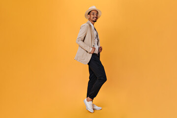 Fototapeta na wymiar Stylish guy in black trousers and beige jacket posing on orange background. Young man in suit and hat looking into camera on isolated