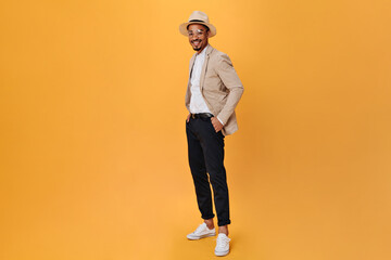 Attractive man in hat and glasses smiles on orange background. Young guy in beige jacket and black...